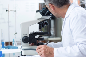 Scientist in a white lab coat examining samples through a compound microscope in a laboratory setting on a desk with notes nearby, conveying research work.