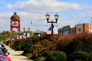 Red clock tower framing a mall plaza with lamp posts, banners, and storefronts along a sunny walkway.