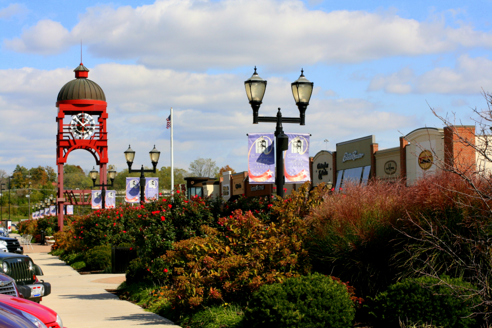 Red clock tower framing a mall plaza with lamp posts, banners, and storefronts along a sunny walkway.