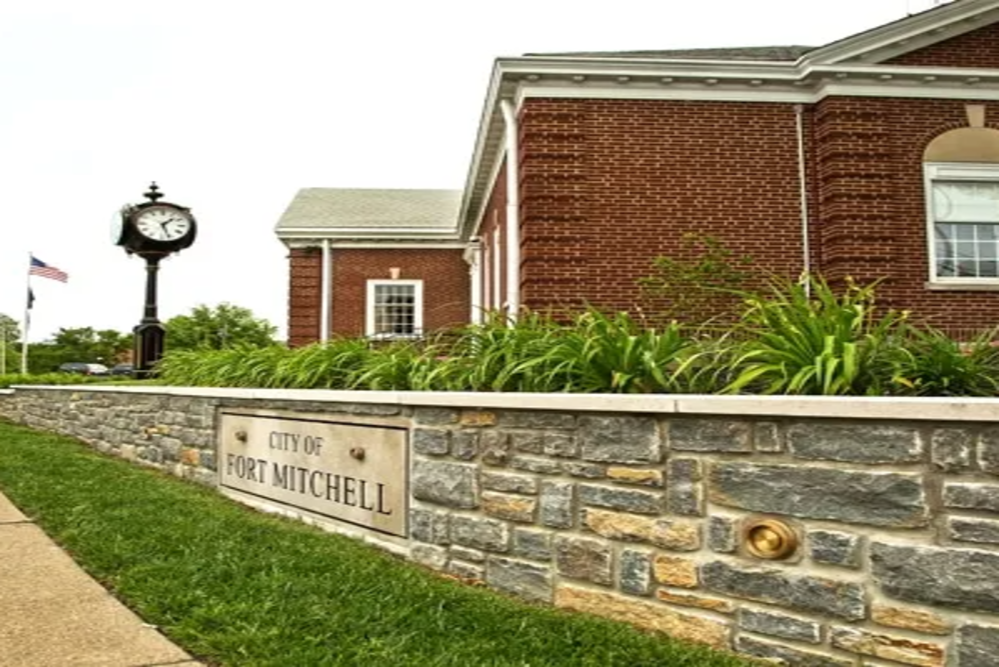 Stone wall with a plaque reading 'City of Fort Mitchell', brick building behind, clock tower to the left, and an American flag in the background.