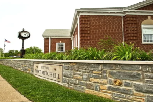 Sign reading 'City of Fort Mitchell' mounted on a stone wall in front of a brick city building with a clock tower and a flag in the background.