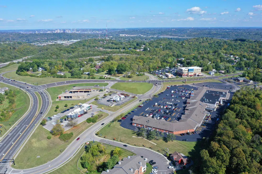 Aerial view of a suburban shopping center with a large parking lot, surrounding roads, and green wooded areas nearby.