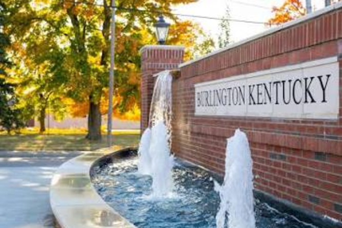 Brick sign reading Burlington Kentucky mounted on a brick wall with a water fountain in front and autumn trees in the background, in fall colors.
