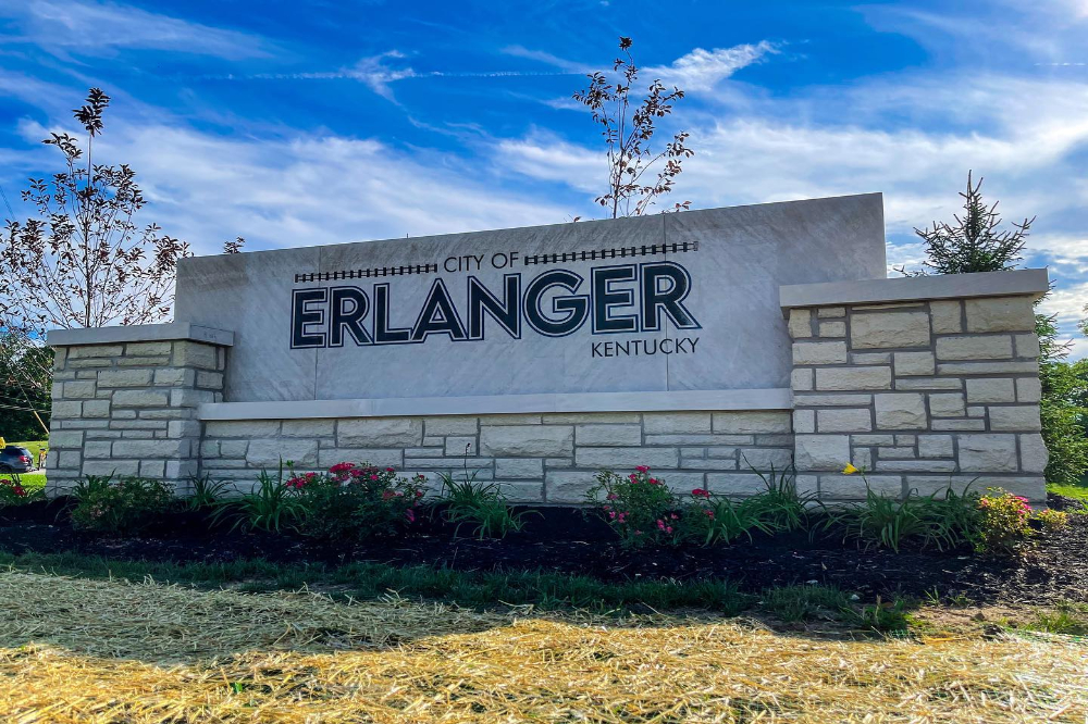 Stone sign reading “City of Erlanger Kentucky” on a light marble backdrop, with a stone planter and flowers in front under a blue sky.