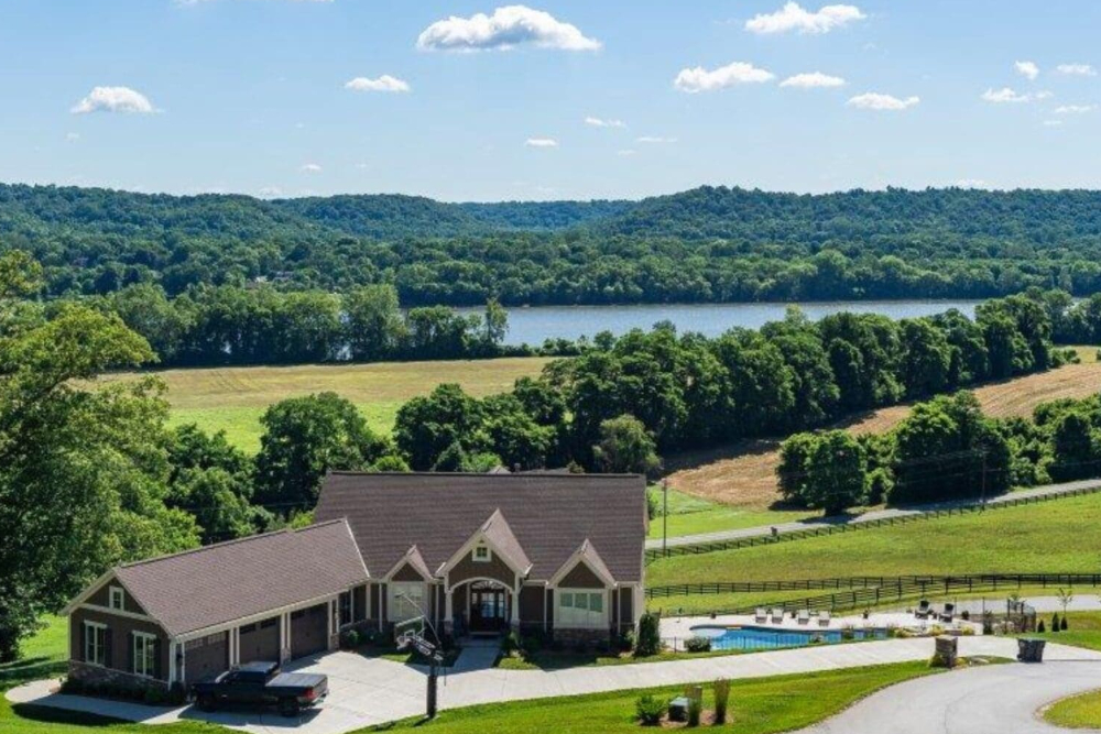 A large house with a dark roof sits in a green, rolling landscape with a pool in the yard and a driveway nearby.